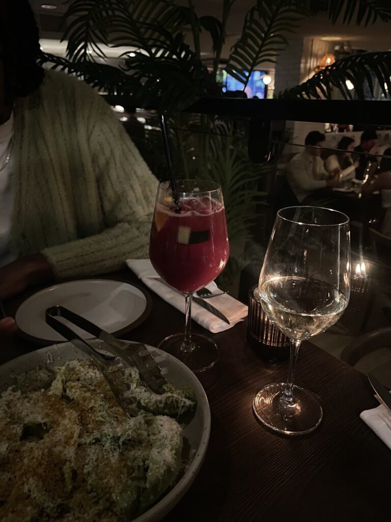 A dimly lit restaurant table with a red sangria, a glass of white wine, and a bowl of greens topped with crispy crumbs.