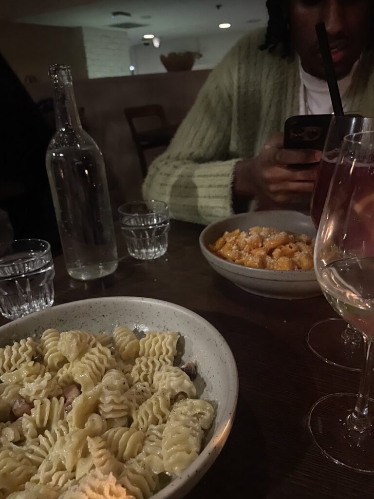 A bowl of creamy rotini pasta in the foreground, with a second pasta bowl, a water bottle, and wine glasses behind it.