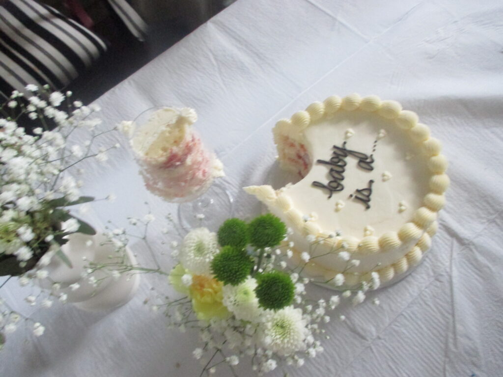 A white buttercream cake with a slice missing, piped with the unfinished words "baby is a...", beside a cut slice in a glass and small white and green flower arrangements on a white tablecloth.