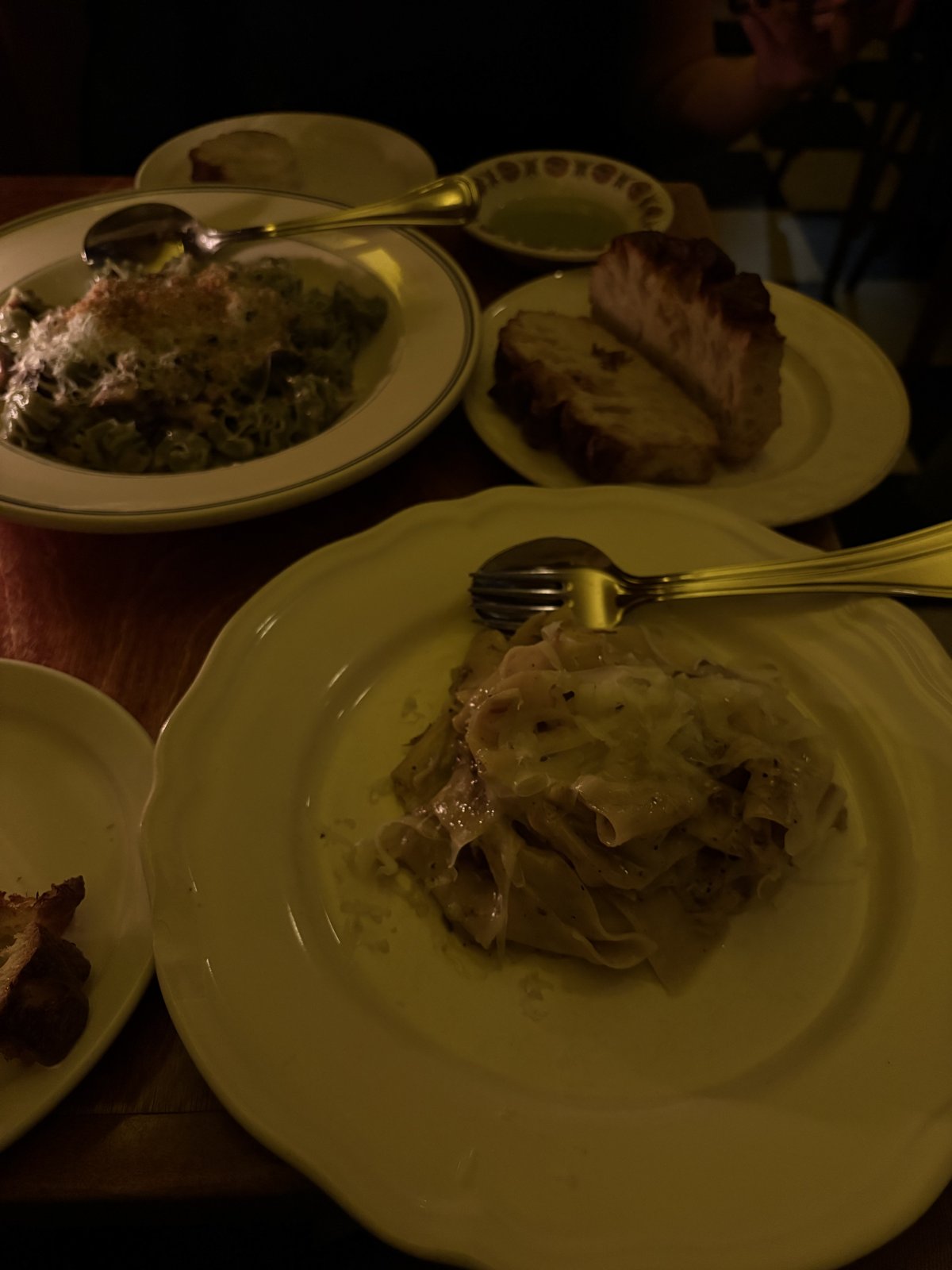 Dimly lit table at Ask for Luigi with a bowl of campanelle with beef ragu, a plate of pappardelle with mushrooms and fonduta, and a plate of focaccia with a small dish of olive oil.