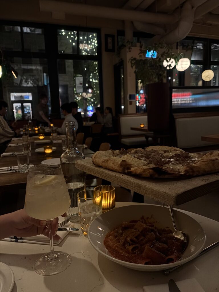 A Primavera pizza on a wooden board above a bowl of rigatoni in tomato sauce, with a lemon spritz in the foreground at a candlelit restaurant in Gastown.