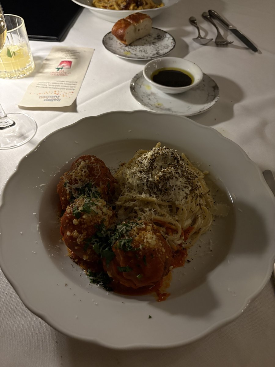 Meatballs in tomato sauce with grated cheese beside a pile of creamy alfredo spaghetti, served in a white bowl at Italian Kitchen, with bread and olive oil in the background.