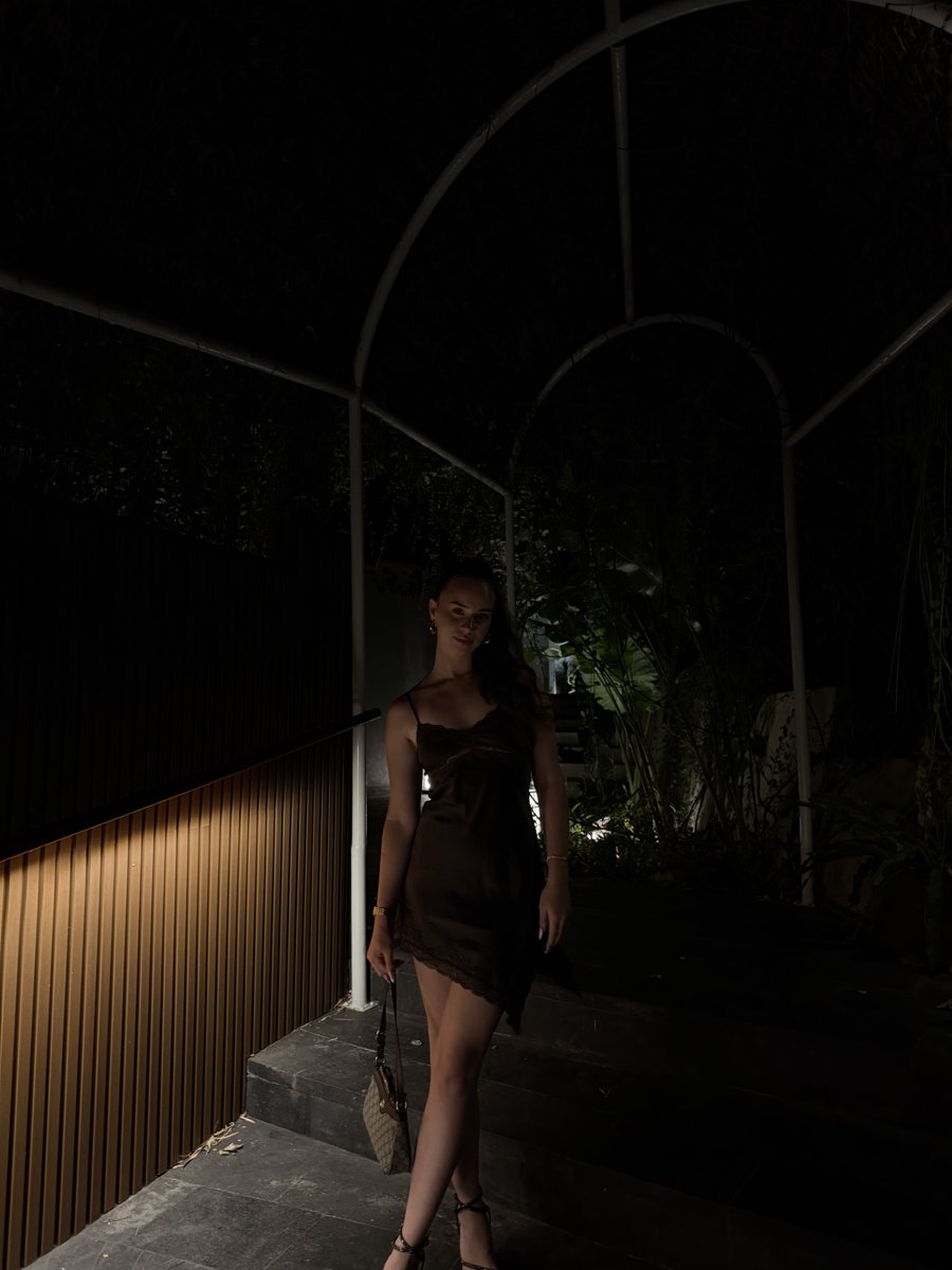 Full-length portrait of Kezjana standing outdoors at night in a brown satin dress, holding a small handbag.