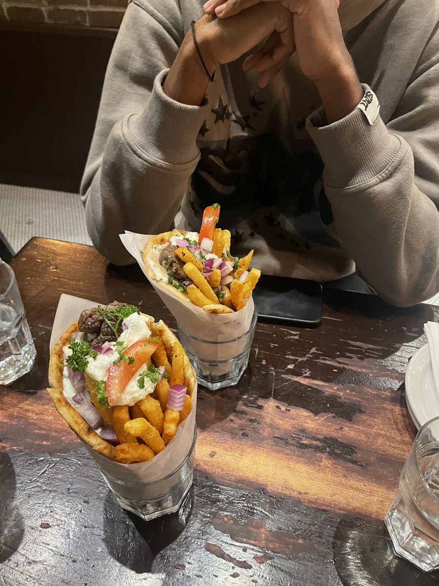Two Greek lamb wraps in paper cones, each filled with fries, tzatziki, tomato, red onion and parsley, on a dark wooden table at Skewers in Gastown.