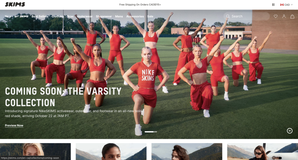 Screenshot of the SKIMS home page showing a large photo of a group of women in red NikeSKIMS activewear posing on a grass field, with the headline Coming Soon: The Varsity Collection over the photo and the menu and search bar across the top.